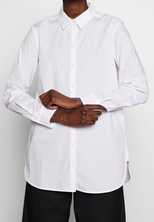 Person wearing a long-sleeve white button-up shirt with a pointed collar, hands crossed at the waist, and black pants.