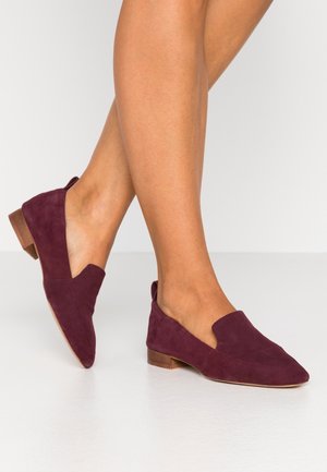 Ben krydset iført bordeaux ruskind slip-on loafers med lave træhæle på en ens hvid gulv og baggrund.