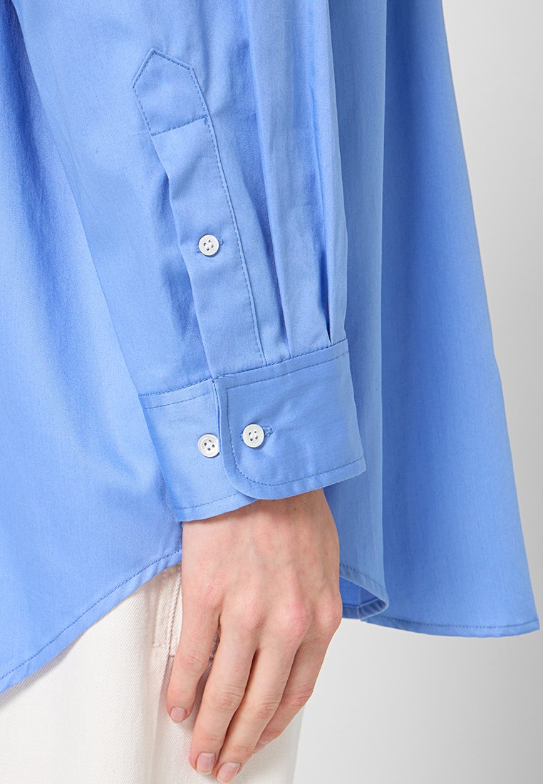 Himmelblaues Langarmhemd mit einem leicht gewellten Saum. Verfügt über Knöpfe an den Ärmelbündchen mit zwei weißen Knöpfen und einer glatten Baumwolltextur.