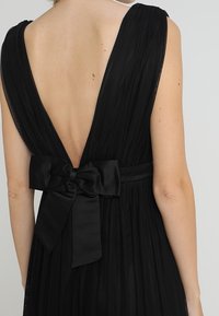 Černé skládané šaty s hlubokým V-výstřihem na zádech a velkou saténovou mašlí v pase. Hladká textura s volným střihem.
