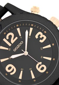 Montre analogique noire avec de grands index dorés, des chiffres proéminents et un bracelet en silicone noir lisse. Comprend une lunette texturée et des boutons poussoirs.