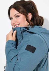 Chaqueta verde azulado con capucha con cremallera, que presenta un parche de logo negro en la manga y cordones de ajuste en el cuello y los puños. Diseño de tela texturizada.
