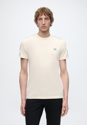 Homme portant un t-shirt crème à manches courtes avec des revers roulés et un petit logo vert sur la poitrine gauche, associé à un pantalon noir, debout devant un fond blanc.