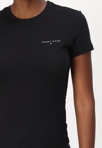 Černé bavlněné tričko s kulatým výstřihem a krátkými rukávy. Na levé straně hrudi se nachází bílé logo "TOMMY JEANS". Hladký povrch.