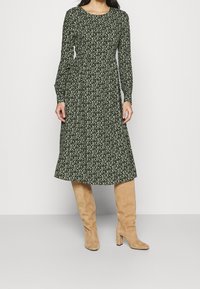 Robe verte à manches longues avec un motif floral, couture à la taille, et jupe évasée. Portée avec des bottes en suède beige jusqu'aux genoux avec un talon carré.