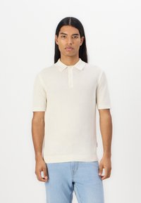 Kremfarget kortermet polo med strukturert strikk, tre knapper foran og ribbestrikkede kanter. Kombinert med lyseblå jeans.