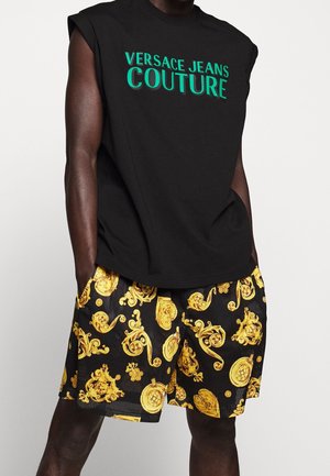 Mężczyzna w czarnej koszulce bez rękawów z zielonym tekstem "Versace Jeans Couture" i czarnych szortach ze złotym barokowym wzorem, ręce w kieszeniach.