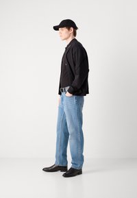 Fekete bomber dzseki egy fekete póló fölött, világoskék egyenes szabású farmer és fekete bokacsizma. Kiegészítők között szerepel egy fekete sapka.