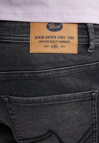 Jeans de mezclilla gris oscuro con una etiqueta de cuero que presenta texto en azul y dorado. Los jeans tienen detalles de costura sutiles y bolsillos estándar.