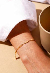 Guldring med ordcharms, der har en delikat kæde og glat finish, båret på et håndled tæt på en neutralfarvet overflade.