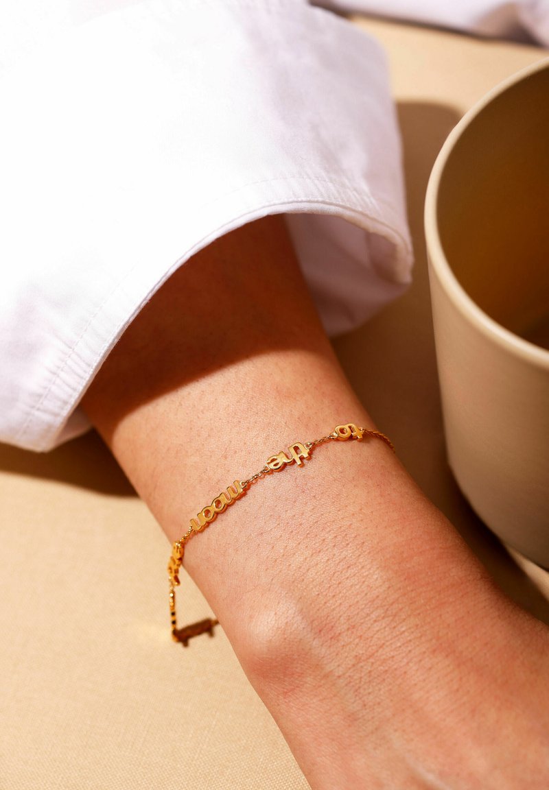 Guldring med ordcharms, der har en delikat kæde og glat finish, båret på et håndled tæt på en neutralfarvet overflade.