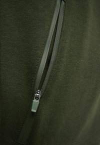 Tejido de punto verde con una cremallera lateral. La cremallera es de color verde oscuro con un tirador de metal, creando una apariencia texturizada.