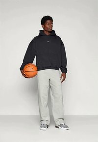 Mann in schwarzem Kapuzenpullover und grauen Sweatpants, der einen orangefarbenen Basketball hält, vor einem einfarbigen hellen Hintergrund stehend.