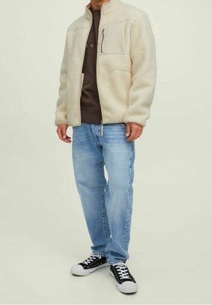 Man in een beige fleecejack, een bruin sweatshirt, een lichtblauwe spijkerbroek en zwart-witte sneakers, staande tegen een effen lichte achtergrond.