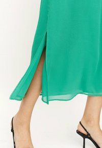 Vestido de gasa verde con una abertura, con una silueta fluida y una textura ligera; combinado con elegantes sandalias de tacón negro.