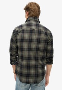 Geruite shirt in donkerblauw, groen en bruin tinten; lange mouwen met opgerolde manchetten; stof lijkt textuur te hebben; voorzien van een kraag en label aan de achterkant.