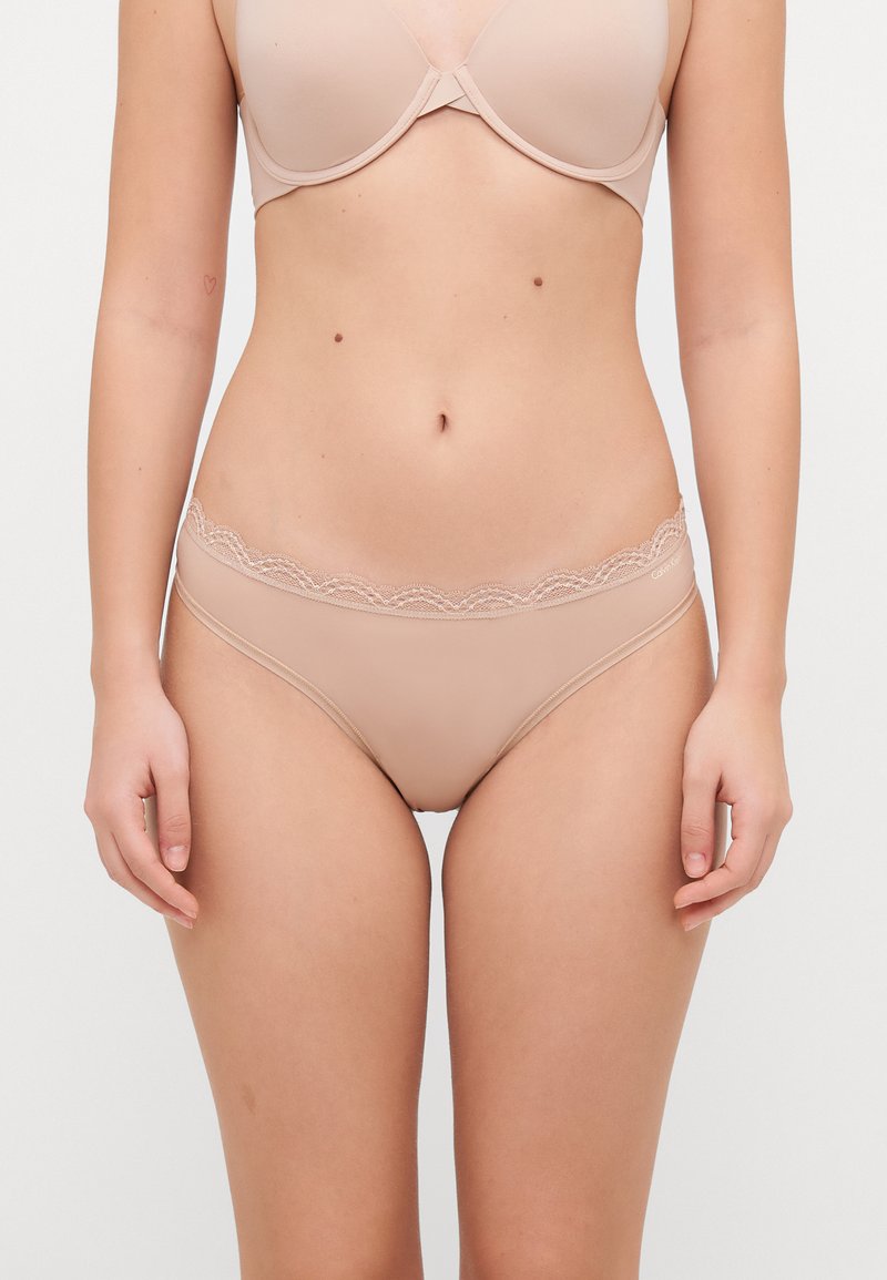 Beige Panties mit Spitzenrand aus glattem Stoff, ausgestattet mit einem gewellten Spitzenbund und einem klassischen Schnitt. Weicher Stoff, minimale Details.