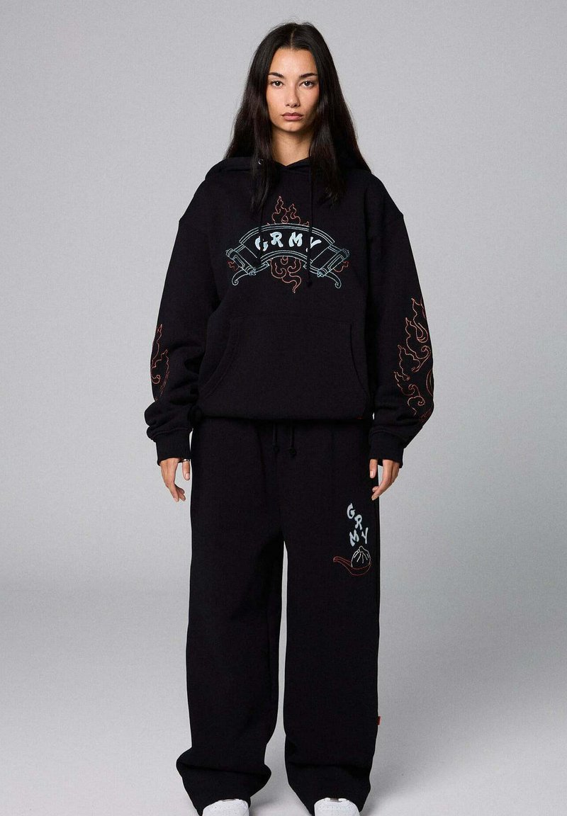 Sudadera negra de gran tamaño y pantalones de chándal, con gráficos bordados en azul y rojo. Tejido suave con un ajuste relajado y un prominente bolsillo canguro.