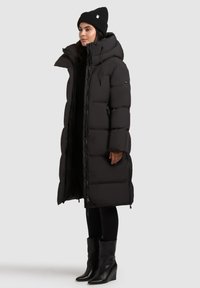 Lang sort pufferjakke med høj krave, der har en frontlynlås, knapdetaljer og en let skræddersyet pasform over sorte leggings og støvler.