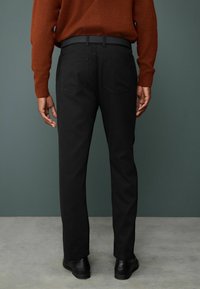 Schwarze Hosen mit geradem Schnitt, ausgestattet mit einem Gürtel, zwei aufgesetzten Taschen und einer glatten Textur. Getragen mit einem rostfarbenen Pullover.