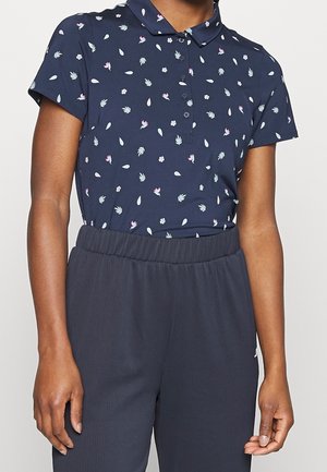 Vrouw die een marineblauw poloshirt met korte mouwen draagt, versierd met kleine bloemenpatronen, en een donker, hooggetailleerd geplooid broek.