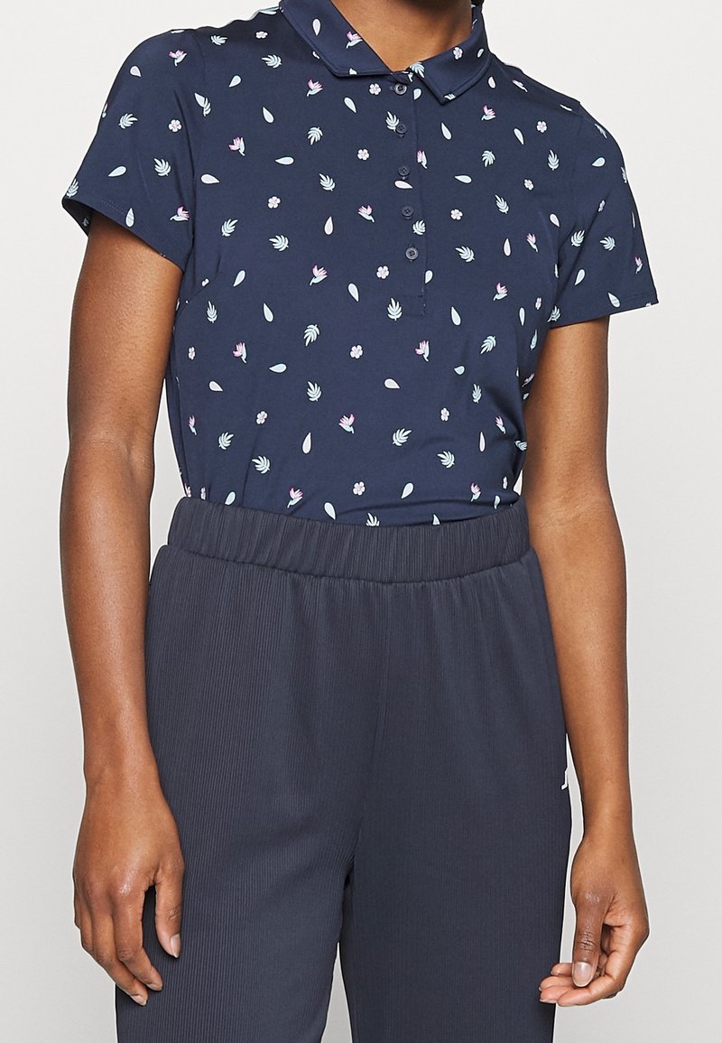 Femme portant un polo bleu marine à manches courtes avec de petits motifs floraux et un pantalon plissé taille haute de couleur foncée.