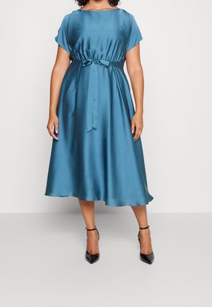 Kobieta ubrana w turkusową satynową sukienkę midi z krótkimi rękawami, paskiem wiązanym w talii oraz czarne czółenka z paskiem wokół kostki i szpiczastym noskiem, stojąca na jednolitym tle.