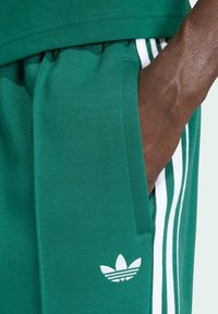 Zelené sportovní šortky vyrobené z měkké tkaniny, s bílými třemi pruhy a bílým logem blízko kapsy. Viditelná hladká textura.