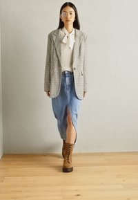 Blazer à carreaux sur un pull blanc côtelé et une jupe midi en denim avec fente avant ; associé à des bottes brunes à lacets. Sol en bois et arrière-plan neutre.