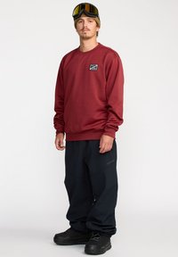 Sweatshirt bordeaux avec des poignets côtelés, associé à un pantalon large bleu marine et des bottes de neige noires. Comprend un patch de logo sur le sweatshirt.