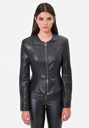 Donna con capelli lunghi e scuri che indossa una giacca di pelle nera aderente con motivi trapuntati e pantaloni di pelle neri su uno sfondo bianco semplice.