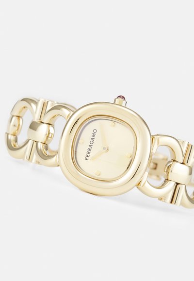 Gold metal watch with a square face, linked bracelet design, and a light yellow dial. Features purple gemstone on the crown and logo detail.