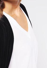 Donna che indossa un top bianco a V sotto un giubbotto nero, mostrata dalle spalle alla parte superiore del torace su uno sfondo neutro.