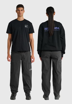 Man die naar voren kijkt draagt een zwart T-shirt met korte mouwen en donkere cargobroek; vrouw met paardenstaart kijkt naar achteren, gekleed in een zwarte trui en bijpassende broek.