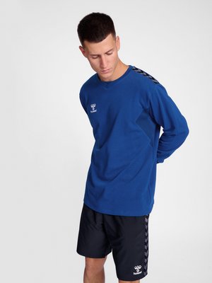 Homme portant un maillot de sport bleu à manches longues et un short noir avec des logos Hummel, regardant vers le bas, les mains derrière le dos sur fond gris.