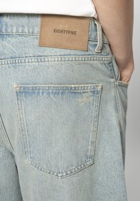Light blue denim jeans with a textured surface, featuring a tan leather patch labeled "EIGHTYFIVE" on the back. Two back pockets visible.