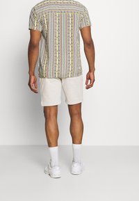 Uomo che indossa una camicia a maniche corte con un motivo a righe verticali e pantaloni corti color beige chiaro, calzini bianchi e sneaker bianche, in piedi di spalle.