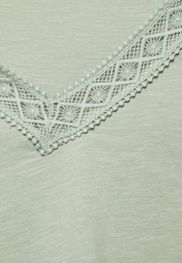 Tecido verde claro com um acabamento texturizado, apresentando uma renda decorativa na gola em V, adornada com padrões circulares e contas.