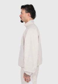 Uomo con capelli ricci e tatuaggi che indossa una giacca e pantaloni chiari, in piedi di profilo contro uno sfondo semplice.