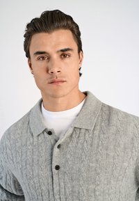 Harmaa neulepusero, jossa on ribbikuvio, kauluksellinen kaula, kolme mustaa nappia ja se on kerrostettu valkoisen paidan päälle.