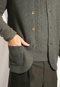 Šedý pletený svetr s kabelovým vzorem, pěti knoflíky a kapsou. Nositý přes žebrovaný svetr a tmavé kalhoty. Měkká textura.