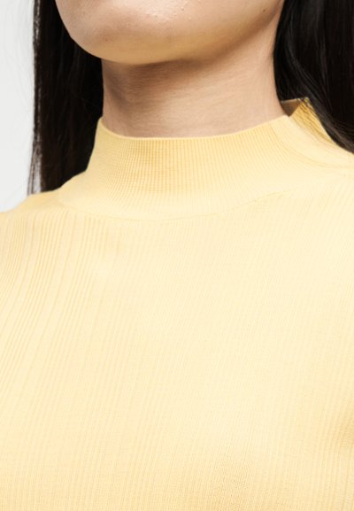 Κοντινό πλάνο του λαιμού και του άνω μέρους του στήθους ενός ατόμου που φοράει ένα ασπριδερό κίτρινο ριμπ πουλόβερ με ψηλό λαιμό, με μακριά σκουρόχρωμα μαλλιά ορατά.
