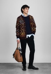 Knit kardigans tumši sarkanā krāsā ar ģeometriskiem rakstiem, uzvilkts virs tumši zila džempera un gaiši zila krekla. Modelis tur brūnu somu ar šalli.