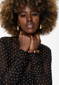 Zwarte doorschijnende top met gouden stippen, lange mouwen en een hoge hals. Het model draagt een dikke amberkleurige en gouden armband en grote gouden oorbellen.