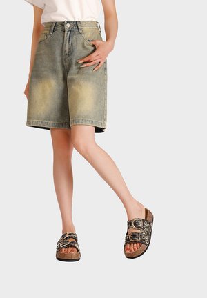 Donna che indossa pantaloncini di jeans verde oliva scoloriti, sandali marroni con doppio cinturino motivo e top bianco con una mano in tasca.