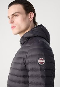 Čierna pufová bunda s kapucňou, s horizontálnym prešívaním a okrúhlym logom Colmar na ľavom pleci. Hladký, lesklý povrch.