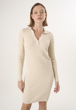 Strickkleid in Creme mit einem Kabelmuster, ausgestattet mit einem kragenlosen V-Ausschnitt, langen Ärmeln und einer figurbetonten Silhouette bis zur Mitte des Oberschenkels.