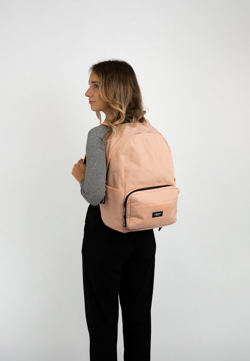 Zaino in morbido tessuto pesca, con un compartimento principale e una tasca anteriore con zip. Il design include spallacci regolabili e una patch con logo.