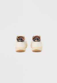 Pantofi sport off-white cu talpă joasă, cu inserții negre în zona călcâiului care afișează logo-ul „Blauer” auriu, tălpi de cauciuc bej, văzuți din spate pe un fundal simplu.