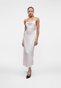 Ψηλή γυναίκα με πλεξούδα, ντυμένη με ένα ανοιχτό ροζ σατέν φόρεμα και μαύρες γόβες με μυτερή μύτη, στέκεται απέναντι σε λευκό φόντο.